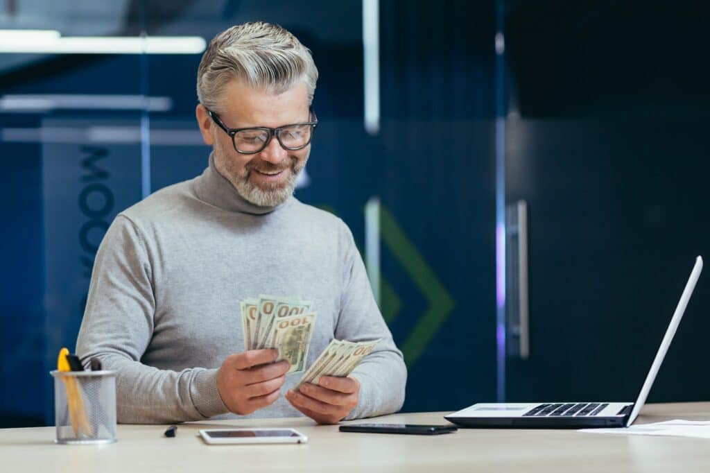 Mature Successful Financier Investor Counts Money Dollars Cash Sitting Inside Office At Table